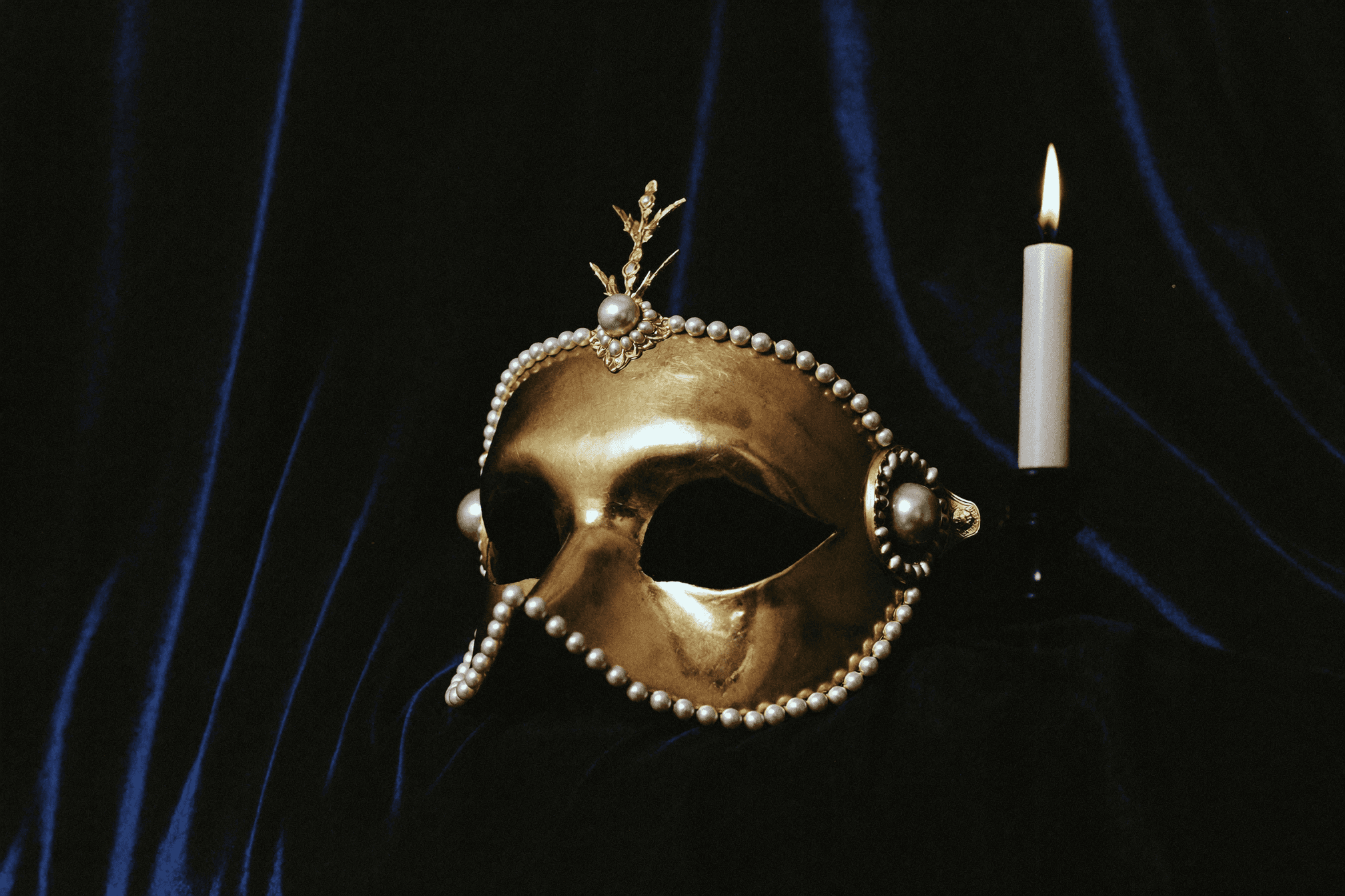 Maschera veneziana in oro antico e perle su velluto blu notte, illuminata da una candela.
