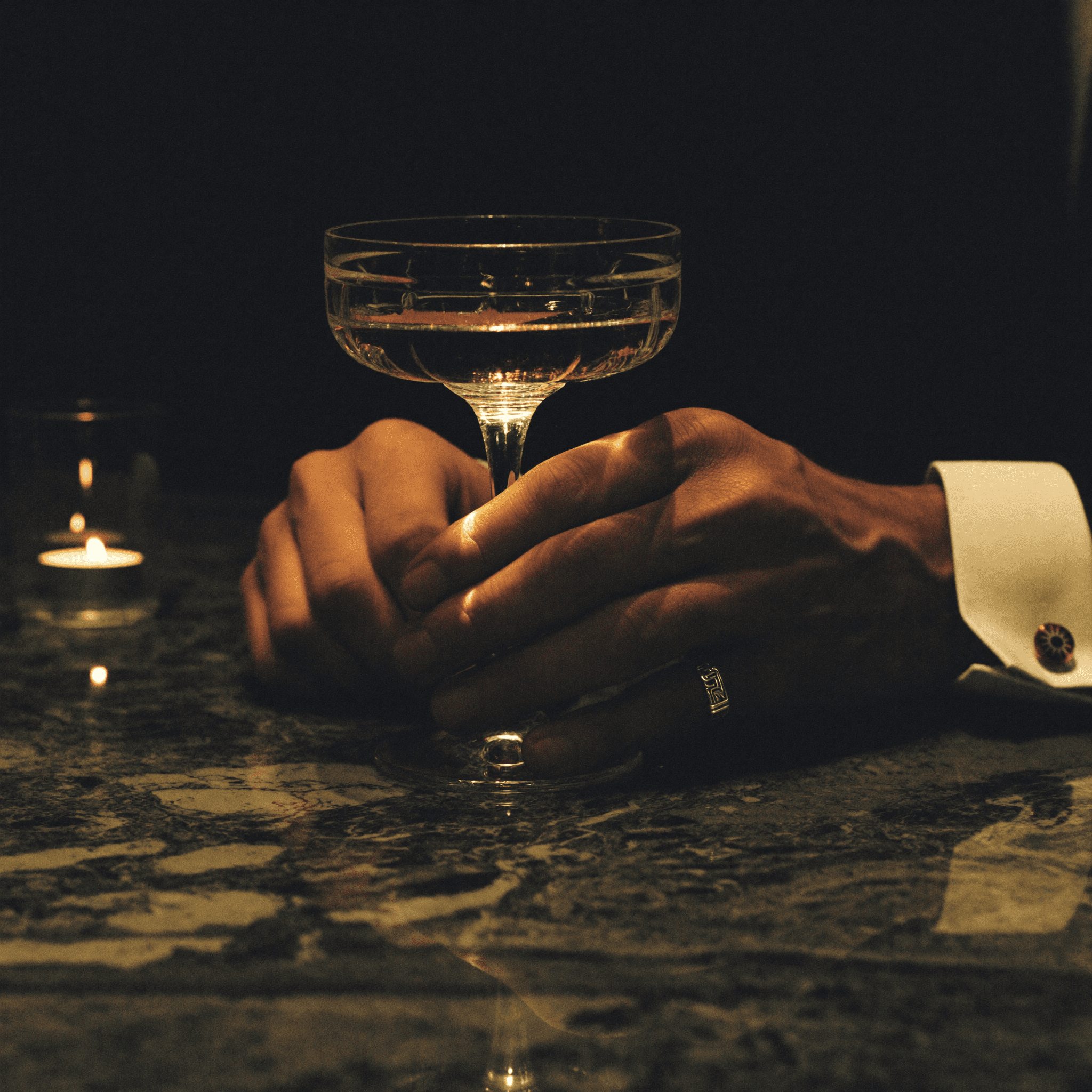 Mani di uomo con anello da mignolo che stringono un calice di cristallo con vermouth ambrato su marmo scuro.