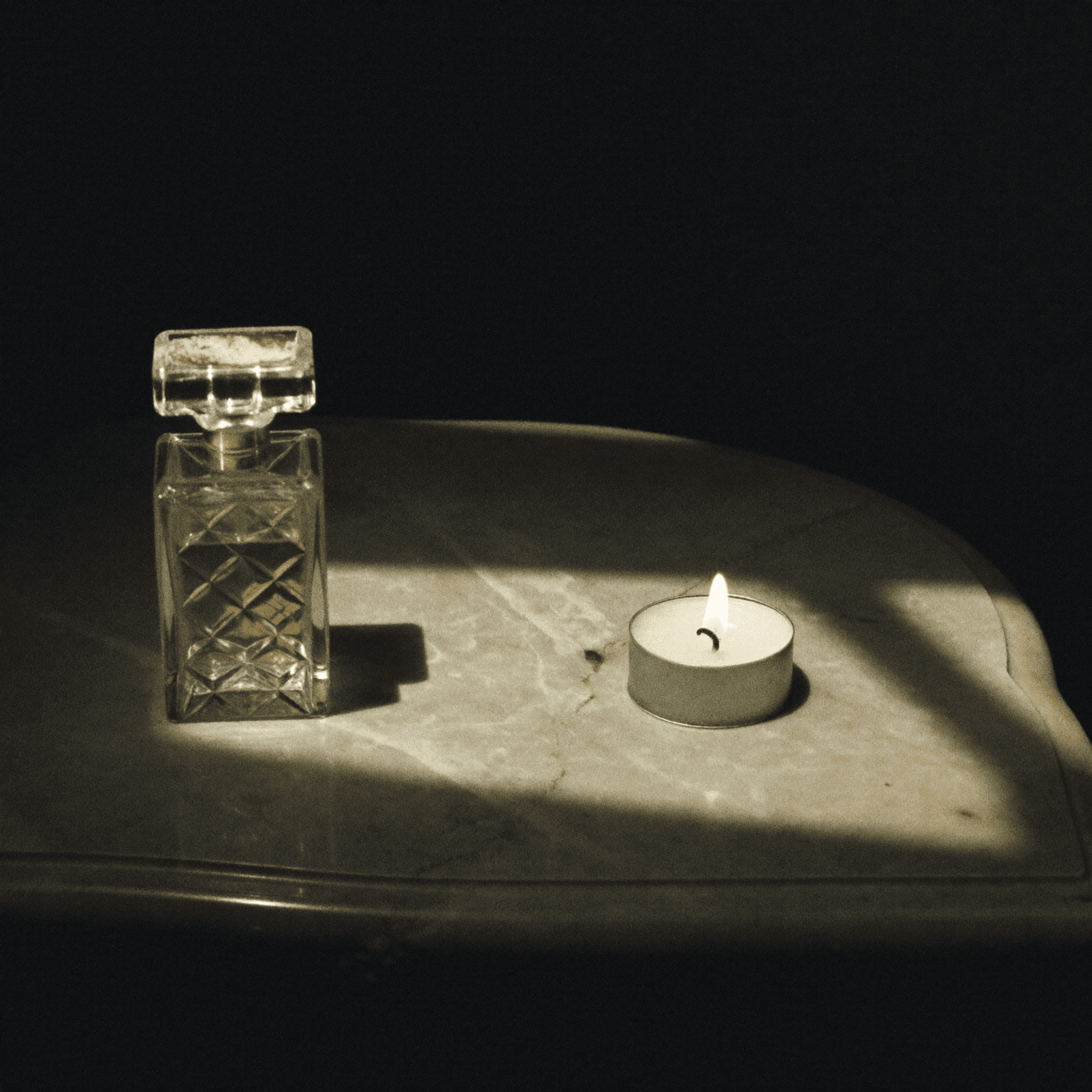 Flacone di profumo in cristallo con tappo dorato su tavolo di marmo, illuminato da un tea light.
