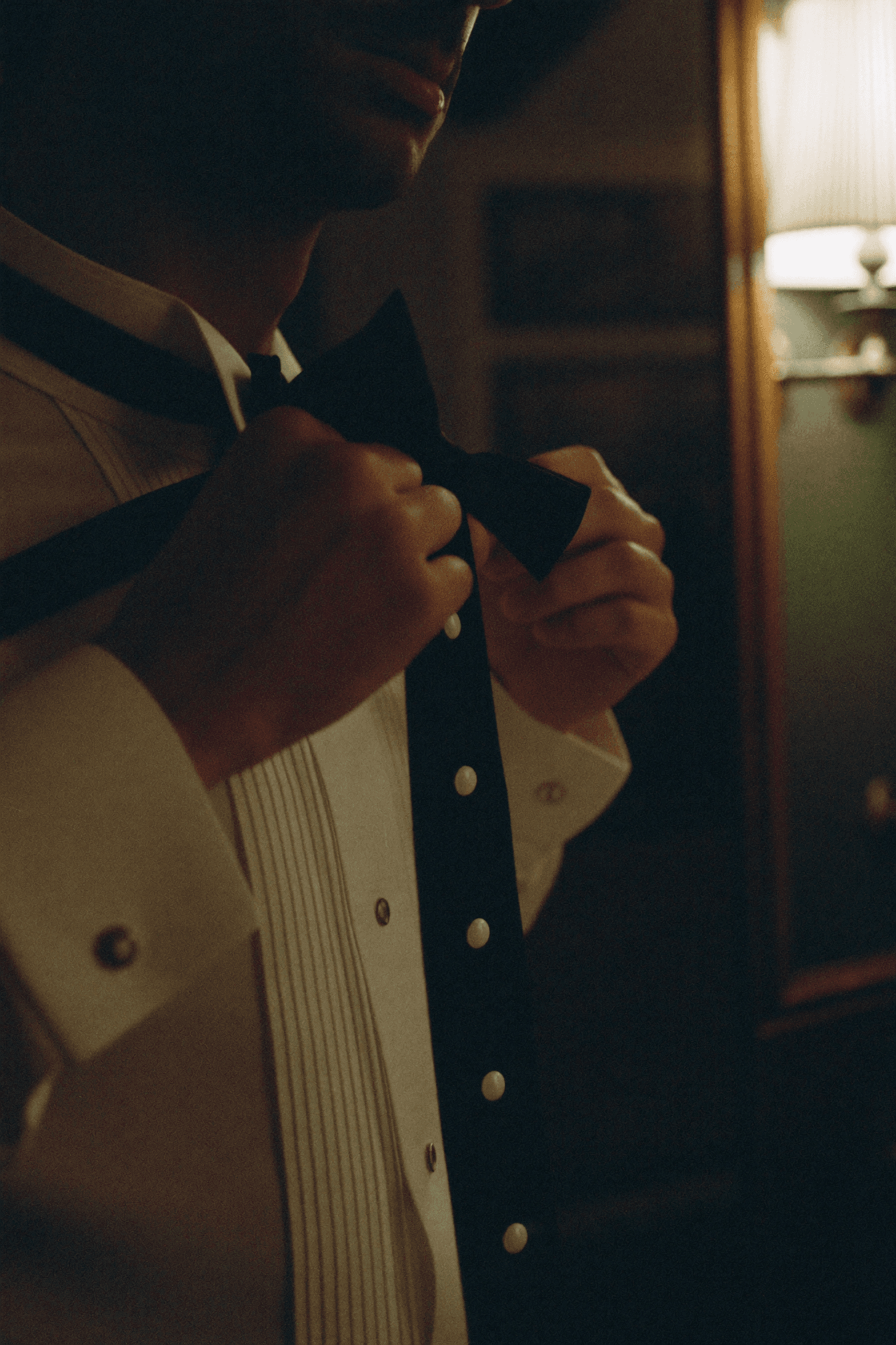 Mani di uomo nell'atto di annodare un papillon nero, camicia con plastron a pieghe e bottoncini di madreperla.