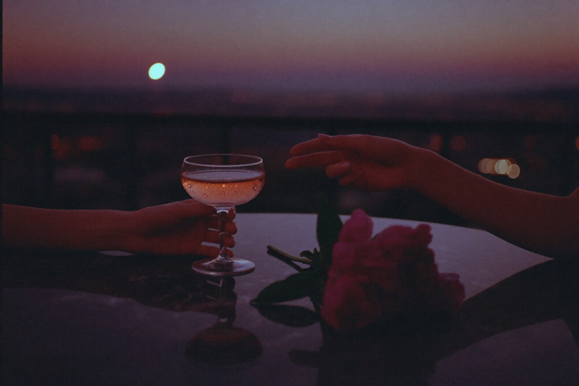 Due mani si incontrano su un tavolo di marmo, una coupe di spumante rosé, una peonia. Terrazza milanese al crepuscolo.